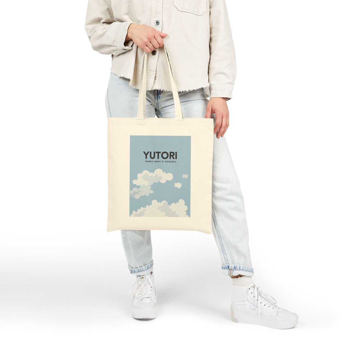 A person wearing a light beige jacket, light wash jeans, and white sneakers holds a cream canvas tote bag with a blue sky and cloud graphic featuring the text 'YUTORI'.