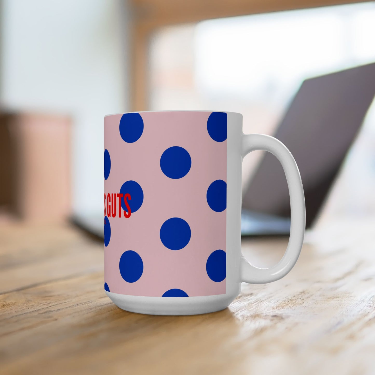 Mug with blue polka dots and red text on a pink background, placed on a wooden surface.