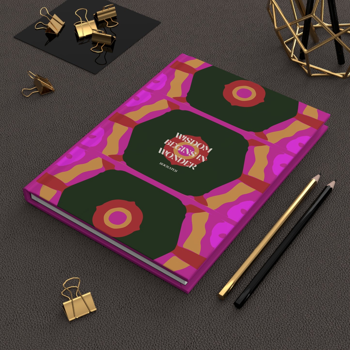 Notebook with geometric design and 'Wisdom Blesses in Wonder' text on a dark surface with office supplies.