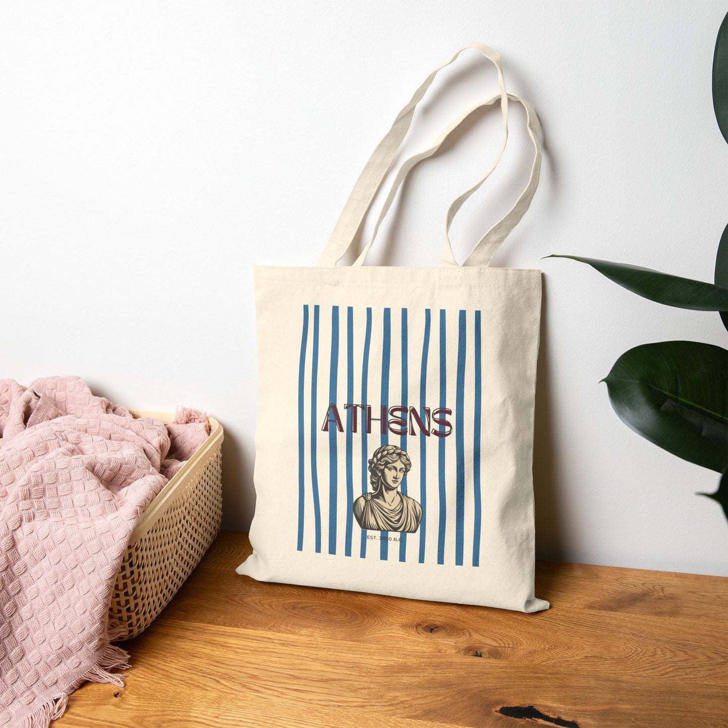 Tote bag with 'Athens' design on a wooden surface with a white wall and plant in the background
