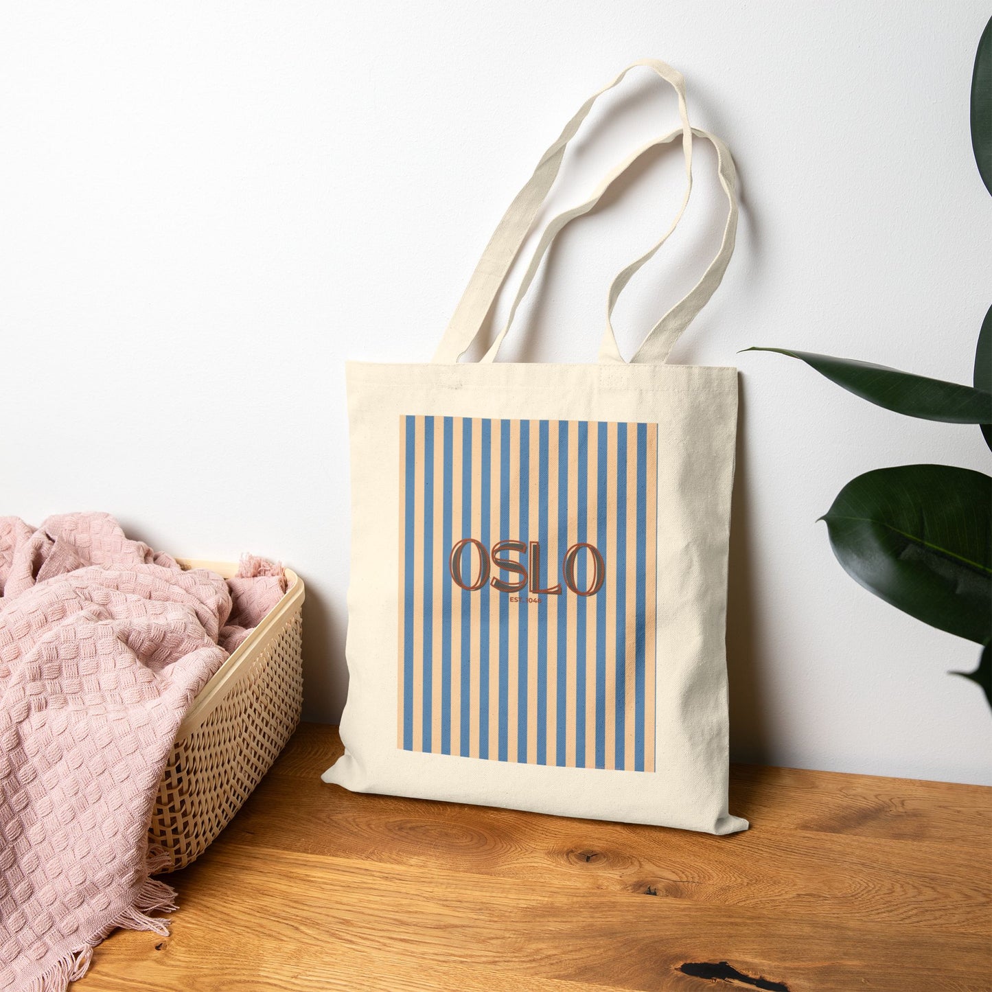 Tote bag with 'OSLO' text and blue stripes on a wooden surface with a plant in the background