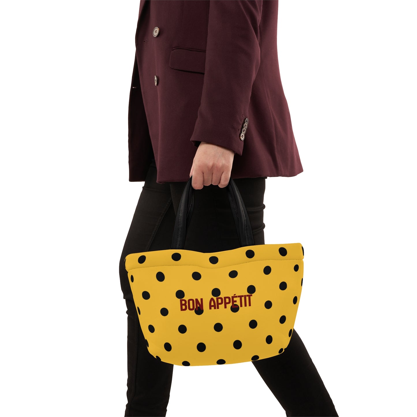 Person holding a yellow polka dot bag with 'Bon Appetit' text, wearing a dark coat.