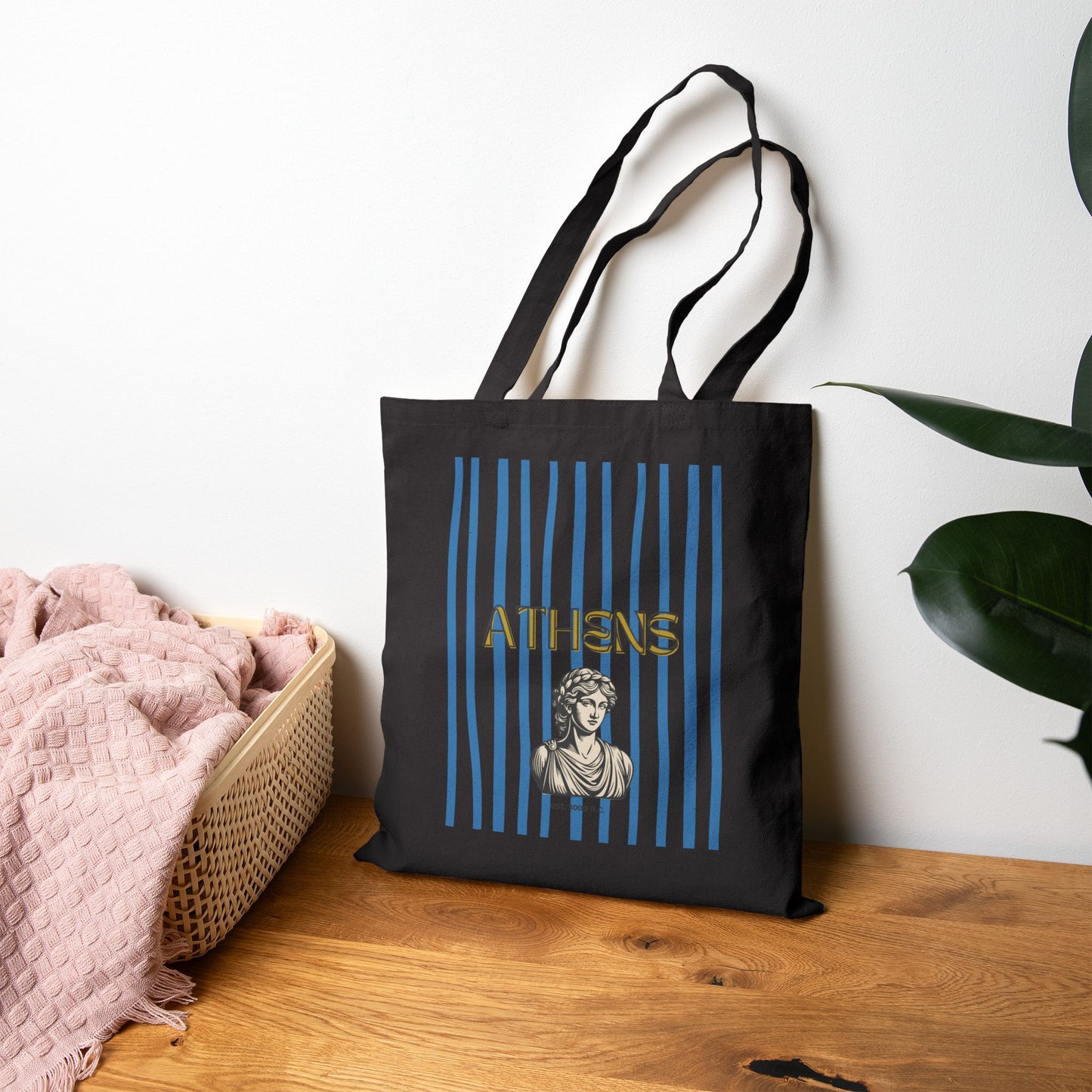 Black tote bag with 'Athens' design on a wooden surface with a plant in the background