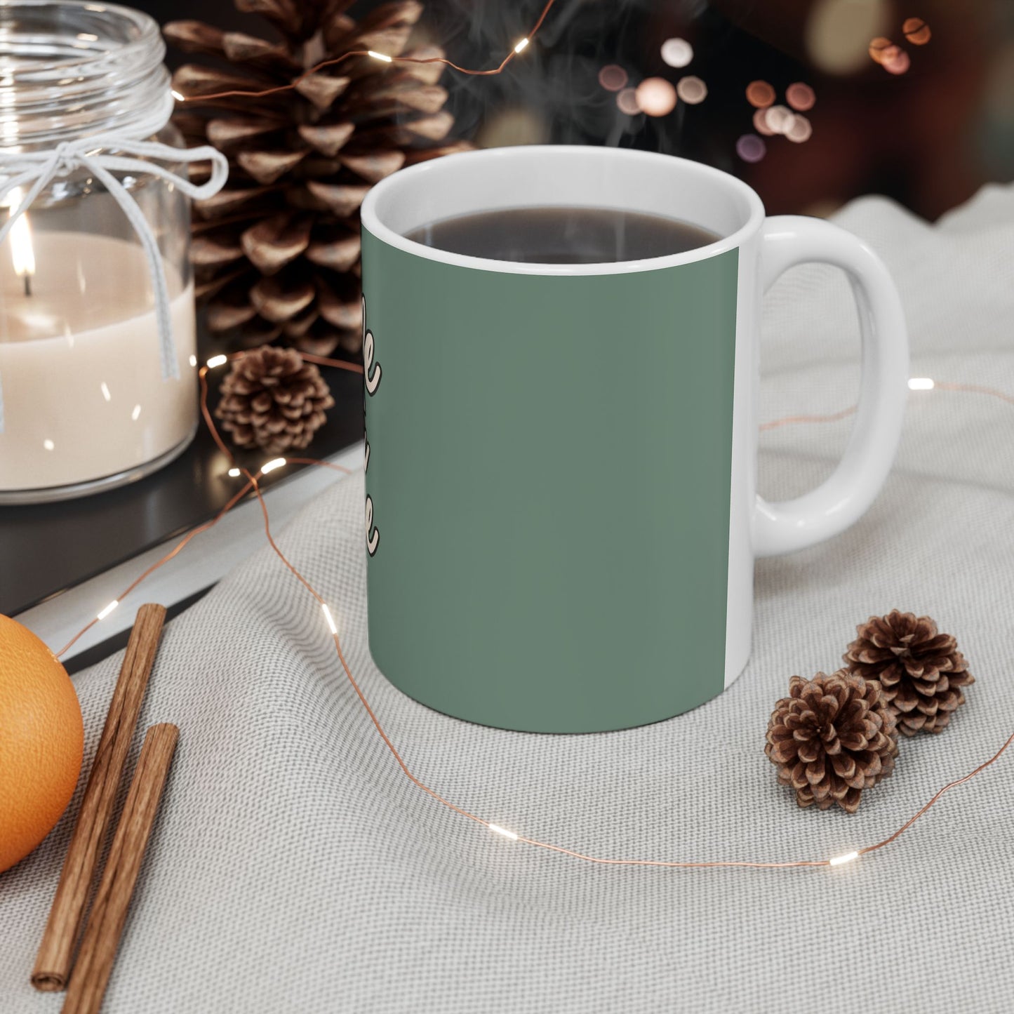 Green mug with steam, surrounded by decorative items like a candle, oranges, and pine cones on a light surface.