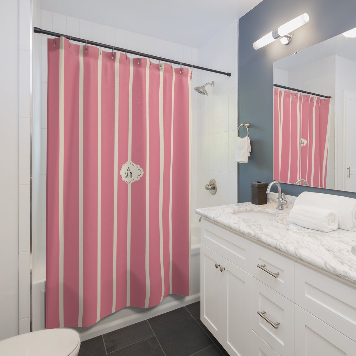 A pink and white vertically striped shower curtain hanging in a modern bathroom with white cabinets, a marble countertop, and a blue-gray accent wall.