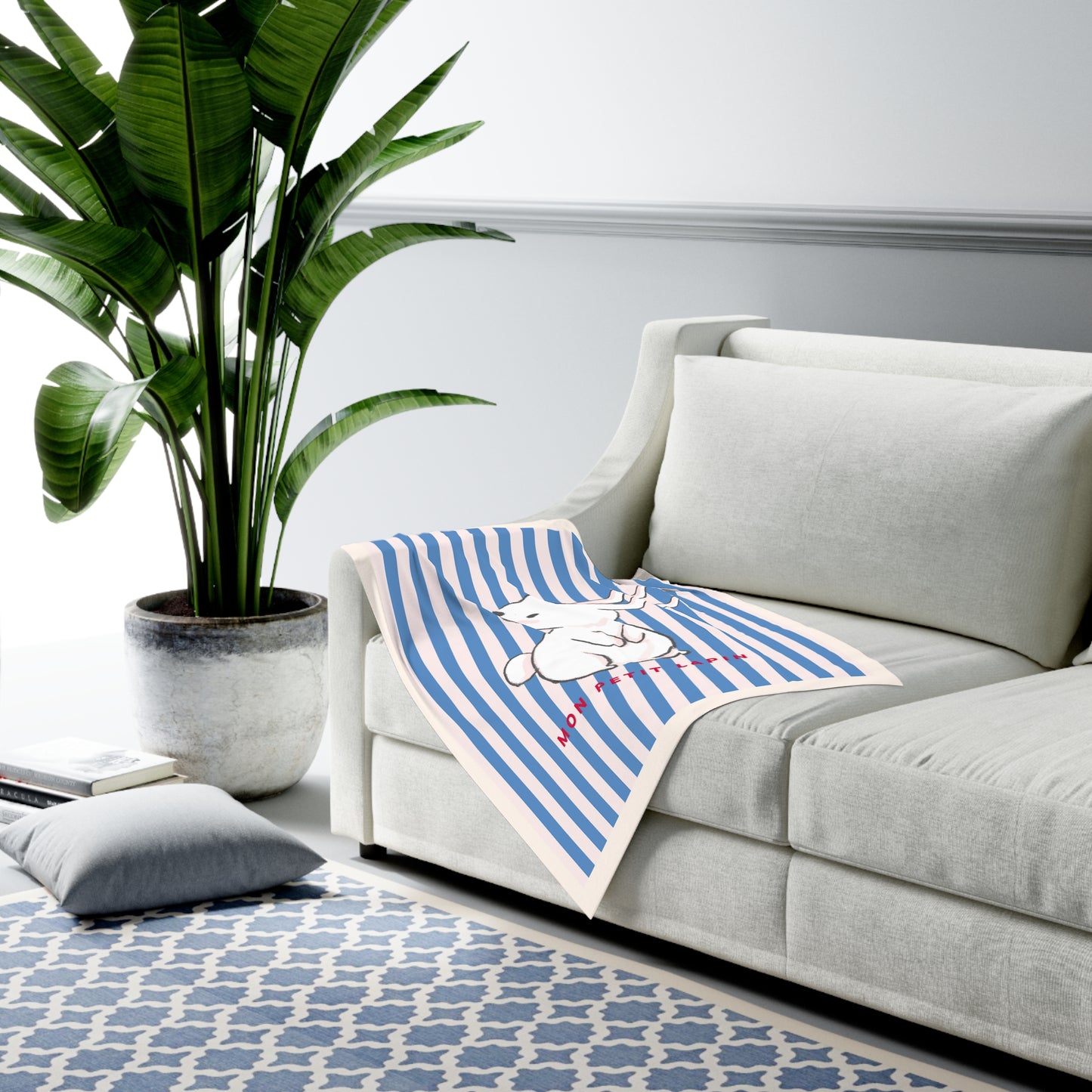 A blue and white striped throw blanket with a cartoon dog graphic is draped over a light gray sofa in a modern living room setting with a potted plant and patterned rug.