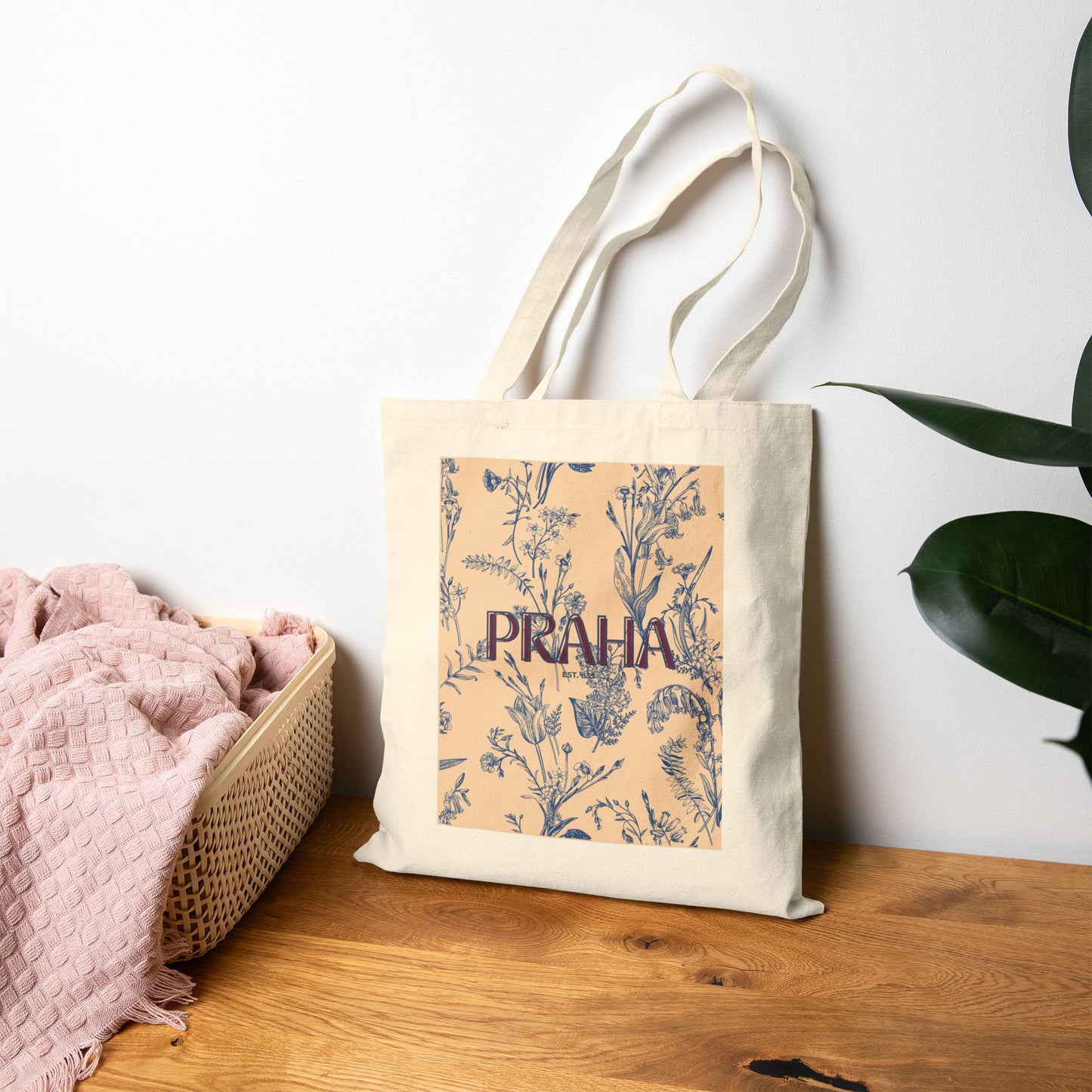 Tote bag with floral design and 'PRAHA' text on a wooden surface with a plant in the background
