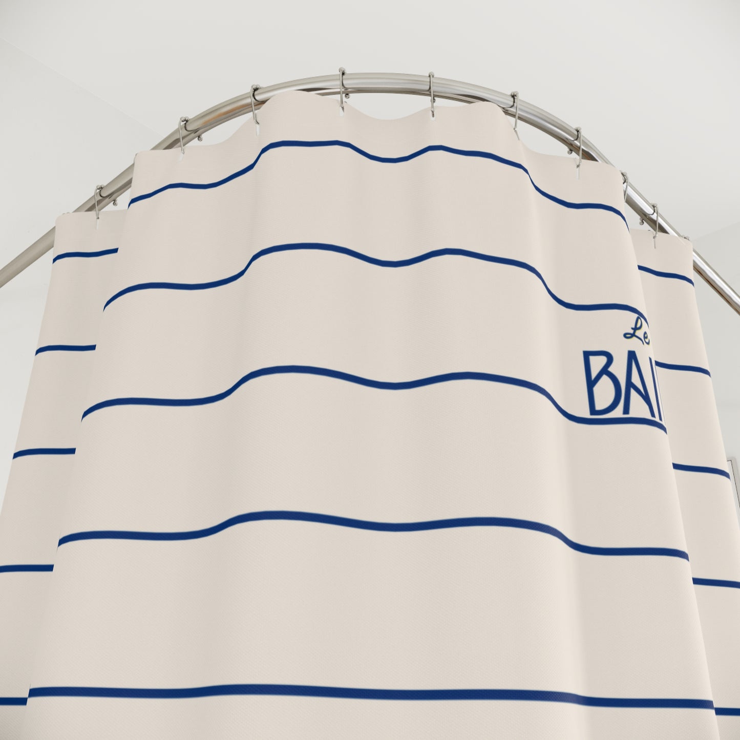 An off-white shower curtain with horizontal navy blue stripes and a navy blue embroidered logo reading 'La Baie' on the right side, hanging from a curved silver metal rod against a light gray wall.
