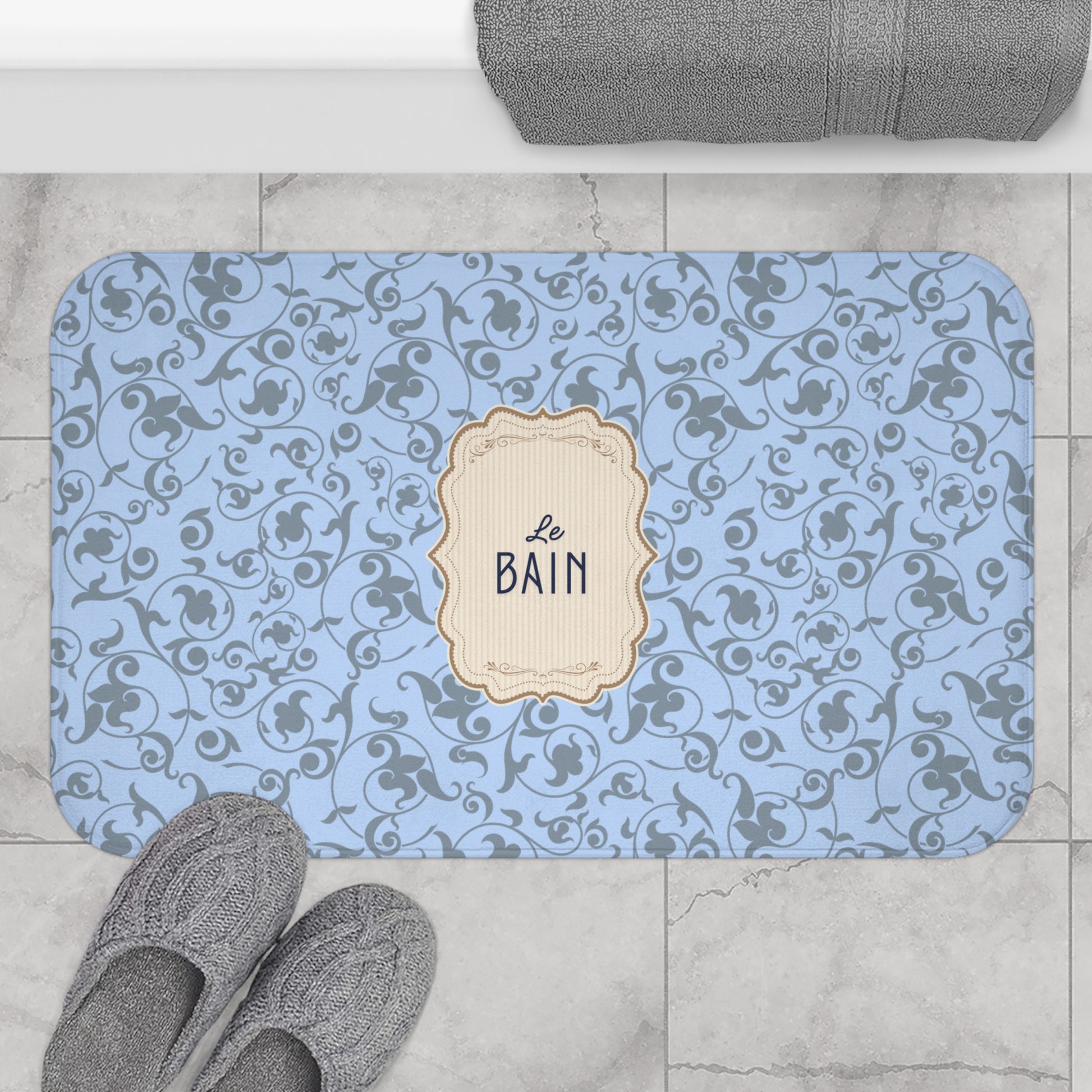 Decorative bath mat with floral pattern and 'Le Bain' label on a tiled floor.