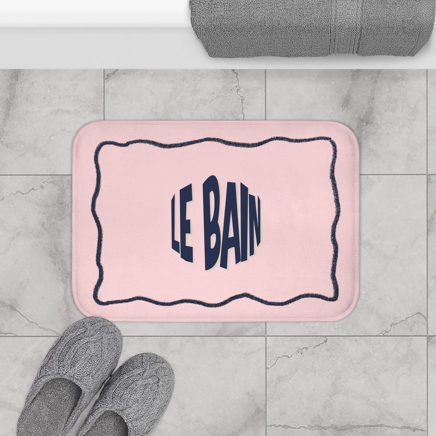 Pink bath mat with 'LE BAIN' text on a tiled floor with gray slippers and a towel.