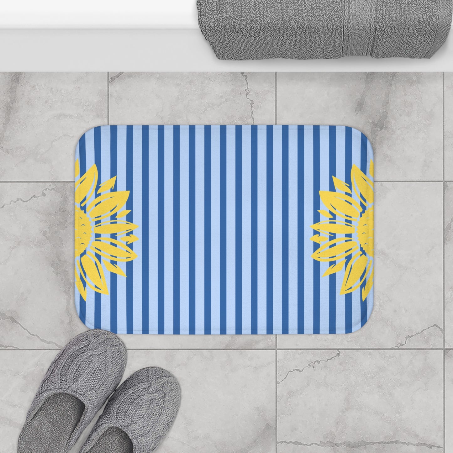 A rectangular bath mat with rounded corners, featuring vertical blue and white stripes with yellow sunflower motifs on both sides, shown on a light gray marble tile floor next to gray slippers and a folded gray towel.