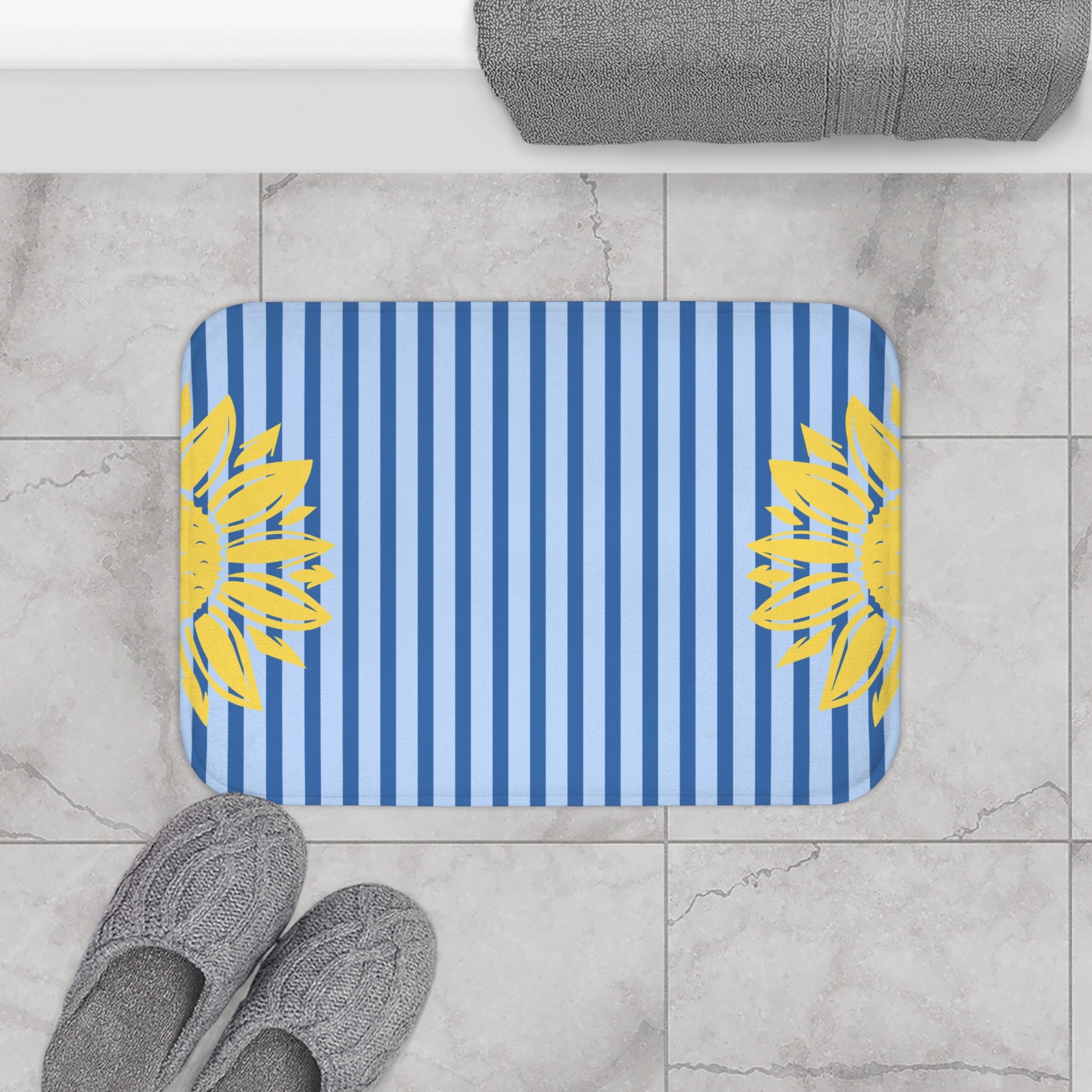 A rectangular bath mat with rounded corners, featuring vertical blue and white stripes with yellow sunflower motifs on both sides, shown on a light gray marble tile floor next to gray slippers and a folded gray towel.