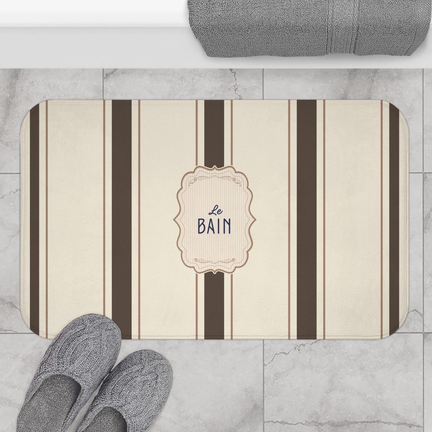 Striped bath mat with 'Le Bain' text on a marble floor with slippers and towel.