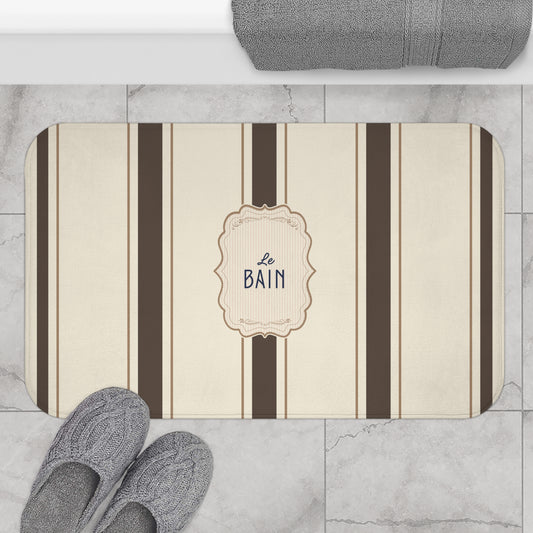 Striped bath mat with 'Le Bain' text on a marble floor with slippers and towel.