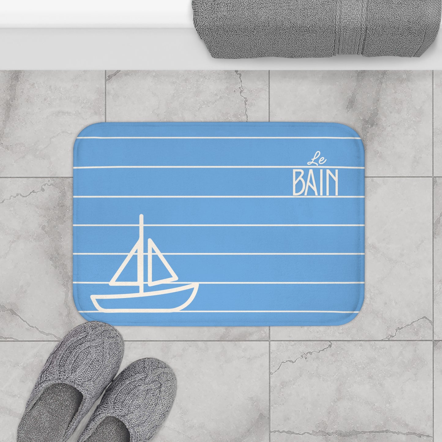 Blue bath mat with sailboat design and 'Le Bain' text on a bathroom floor.