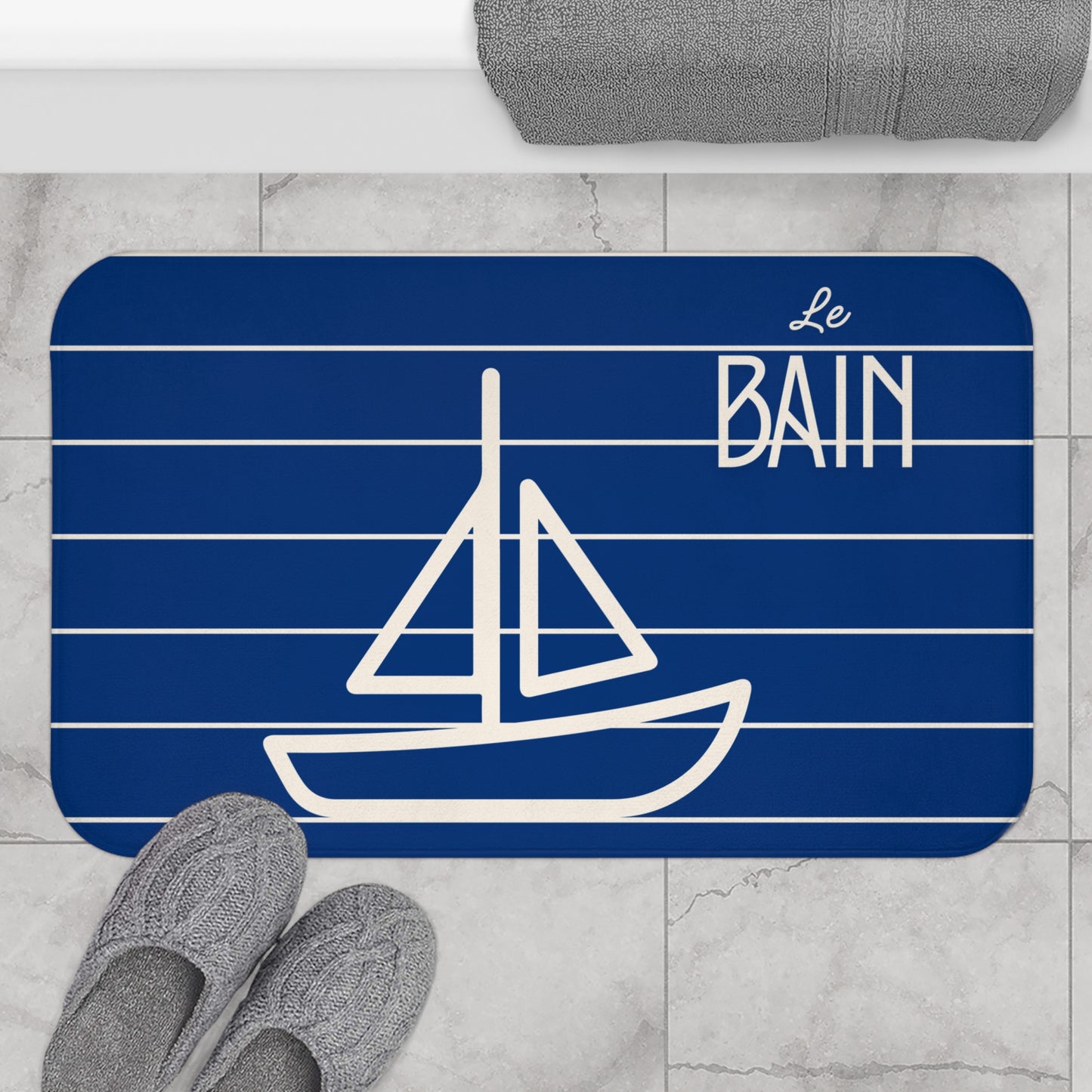 Bath mat with sailboat design and 'Le Bain' text on a blue background, placed on a marble floor.