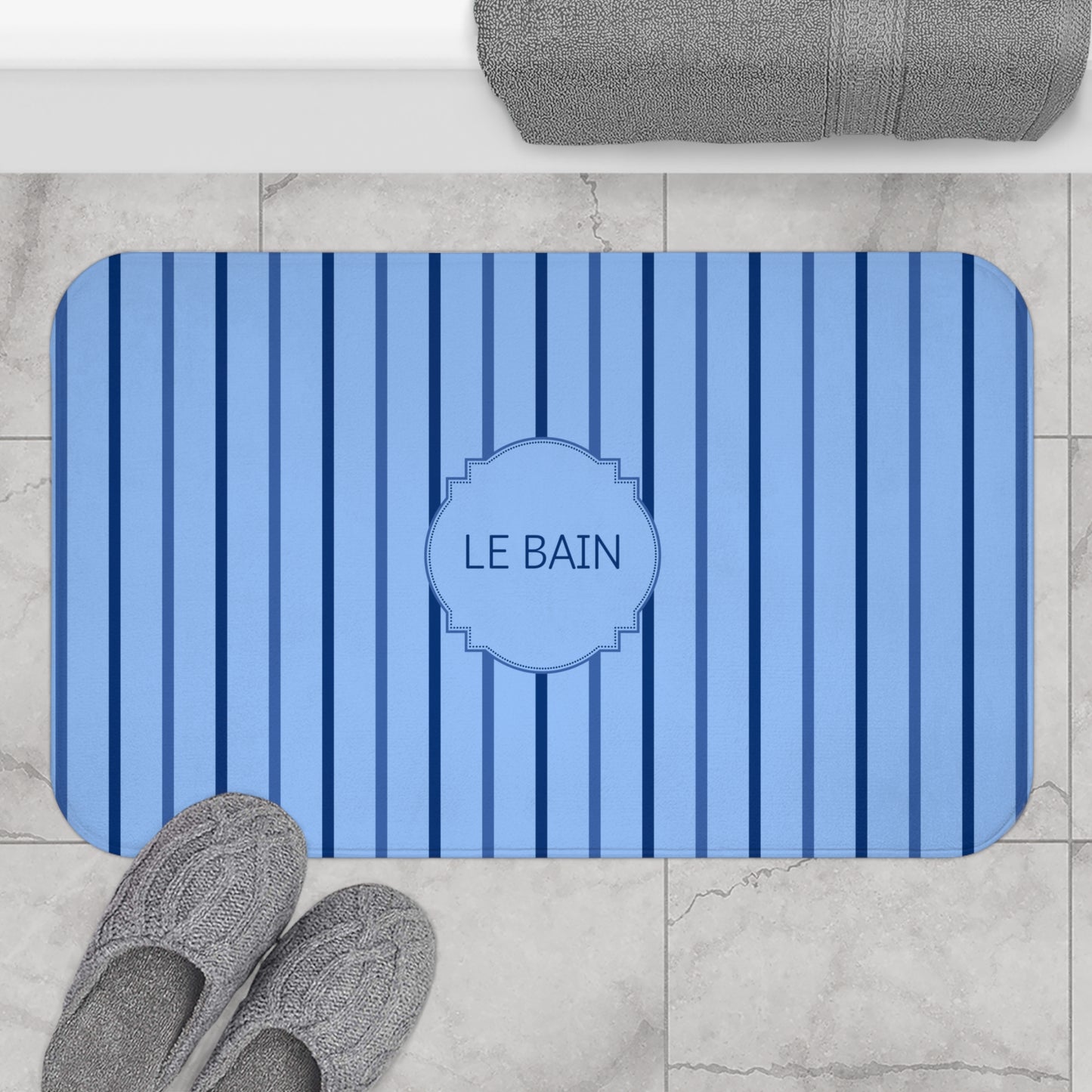 A blue and navy striped bath mat with a central monogrammed label reading 'LE BAIN', shown on a light marble bathroom floor with gray slippers and a folded gray towel nearby.