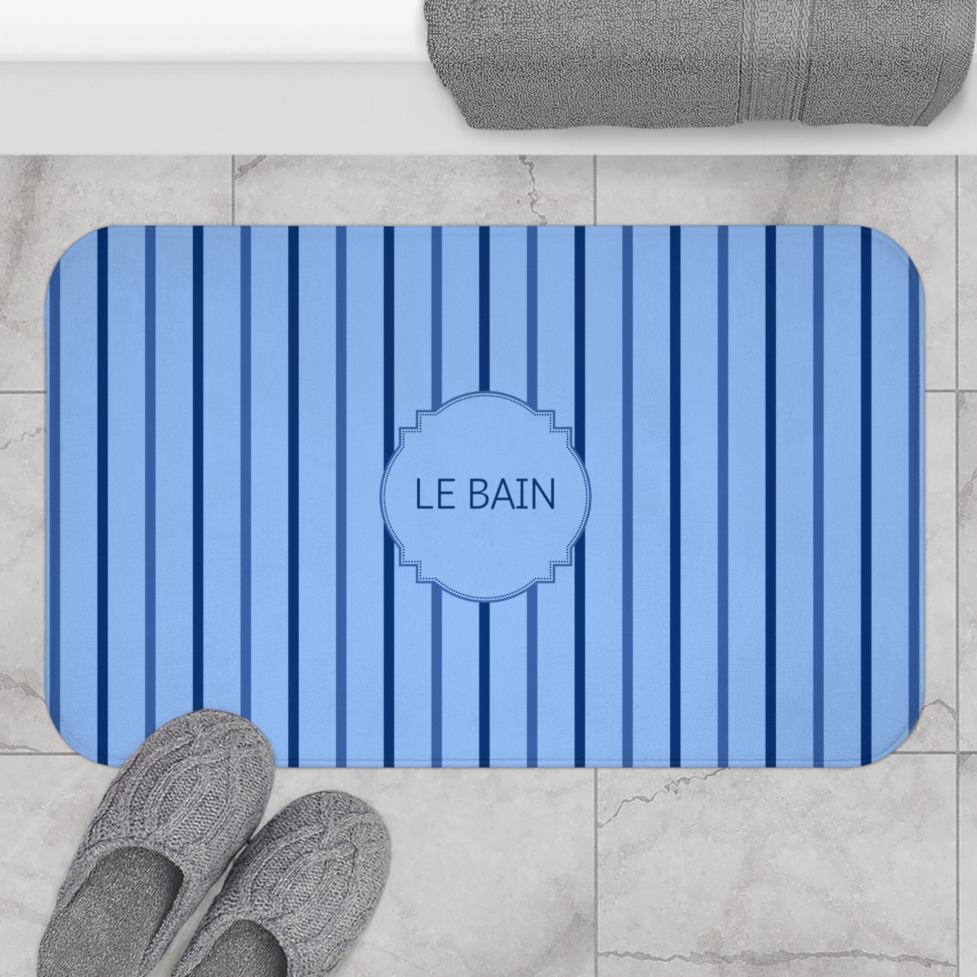 A blue and navy striped bath mat with a central monogrammed label reading 'LE BAIN', shown on a light marble bathroom floor with gray slippers and a folded gray towel nearby.