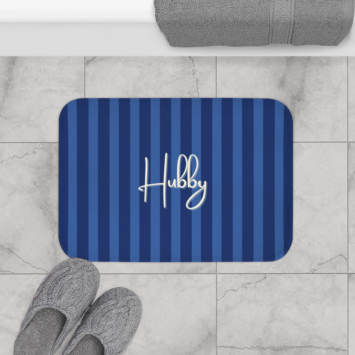 A rectangular bath mat with rounded corners, featuring vertical navy and medium blue stripes and the name 'Hubby' in white script. The mat is placed on a light gray marble tile floor next to gray slippers, with a folded gray towel visible at the top edge.