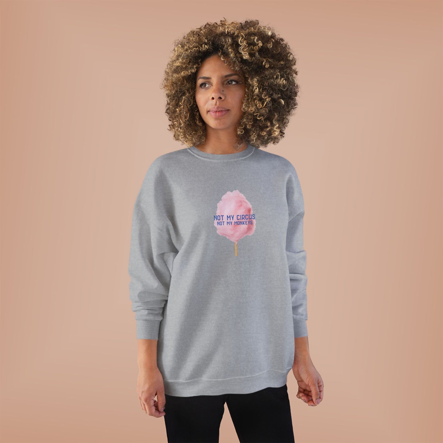A woman with a large curly afro stands against a solid peach studio background wearing a light heather gray sweatshirt with a pink cotton candy graphic and the text 'NOT MY CIRCUS, NOT MY MOKEYS' printed on the chest.