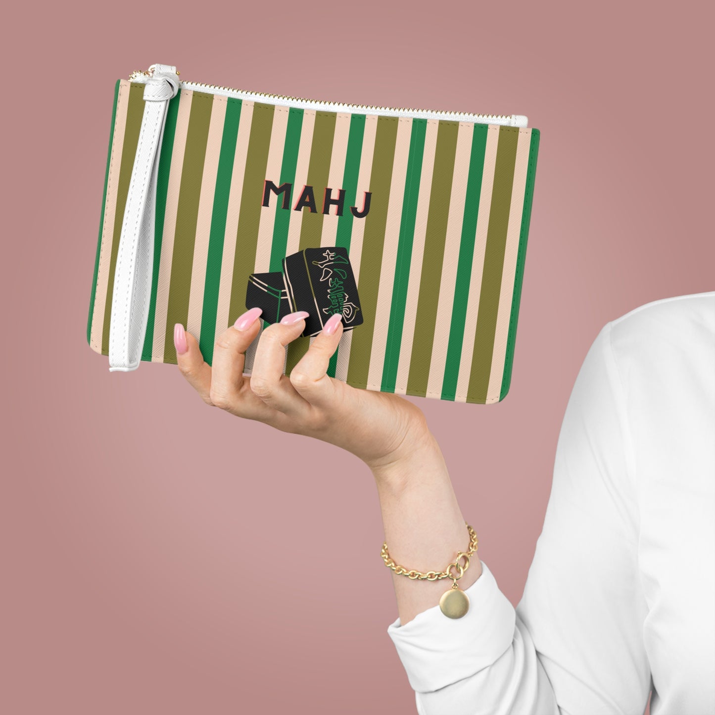 A person holds a zippered pouch with vertical green and beige stripes, a white wrist strap, and the name 'MAHJ' printed on the front. The person is wearing a white long-sleeve top and a gold bracelet, set against a solid dusty pink background.