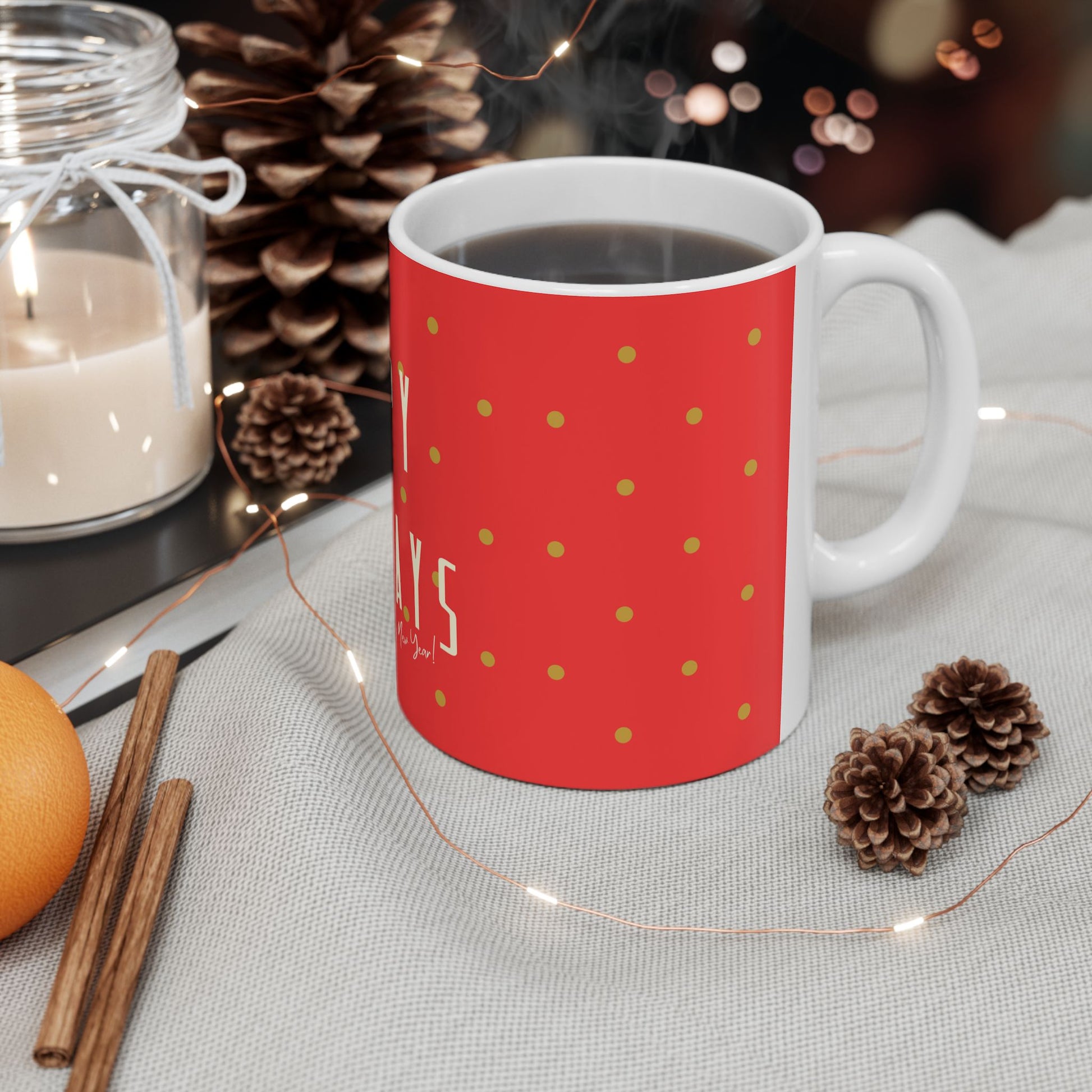 A festive red ceramic mug with gold polka dots and partial white text reading 'MERRY', filled with a hot beverage and set on a light fabric surface with pinecones, fairy lights, a candle jar, cinnamon sticks, and an orange.