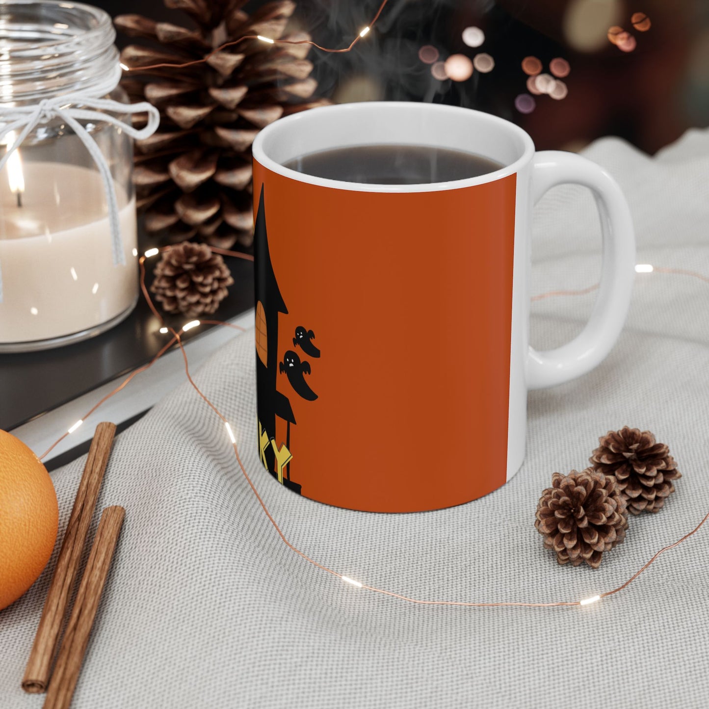 A white ceramic mug filled with a dark beverage, featuring a printed orange Halloween-themed design with a witch's hat and brooms. The mug is styled on a light fabric surface with warm string lights, pinecones, cinnamon sticks, and a lit candle in the background.