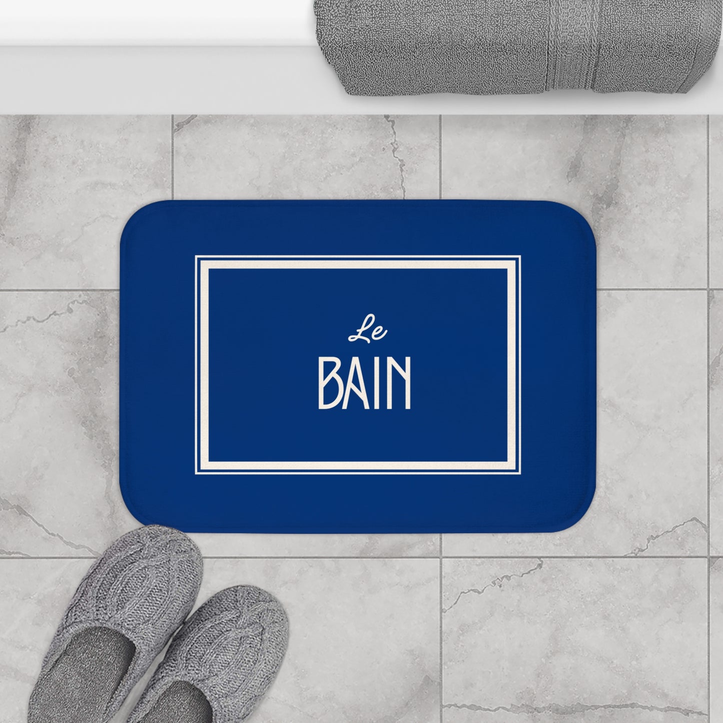Blue bath mat with 'Le Bain' text on a marble floor with slippers and towel.