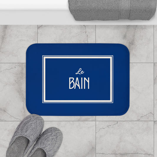 Blue bath mat with 'Le Bain' text on a marble floor with slippers and towel.