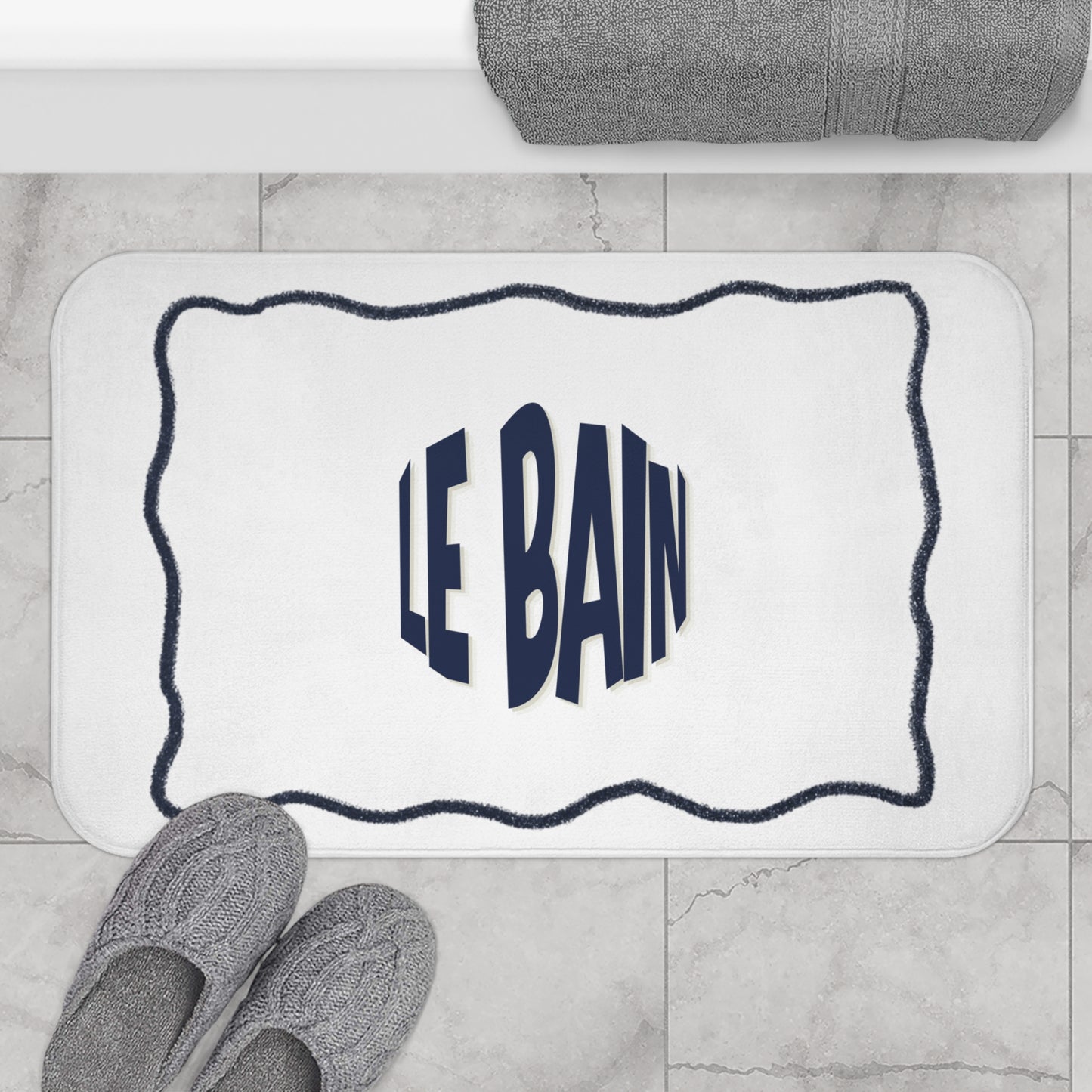Bath mat with 'LE BAIN' text on a bathroom floor with slippers and towel.