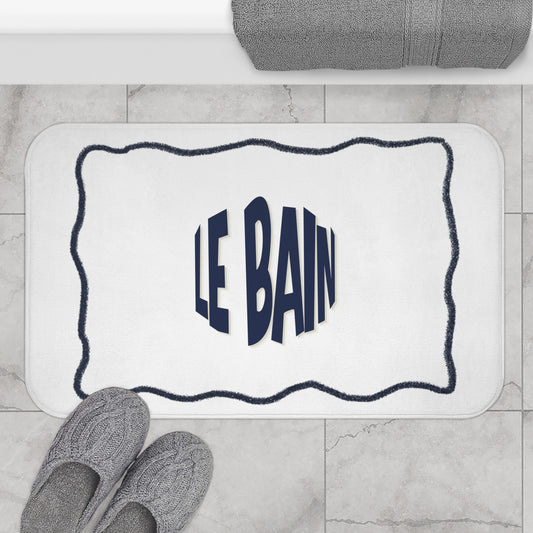 Bath mat with 'LE BAIN' text on a bathroom floor with slippers and towel.