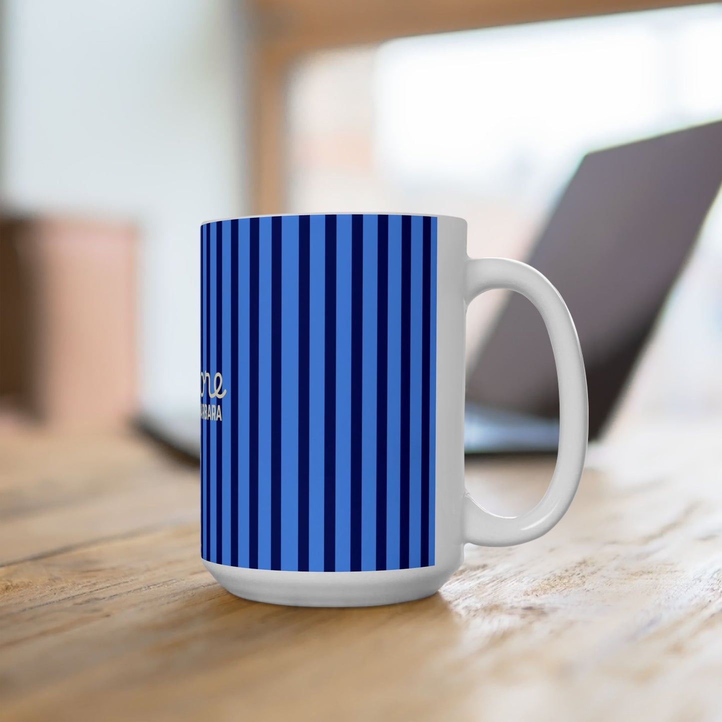 A white ceramic mug with a blue and navy vertical stripe pattern, sitting on a wooden table with a blurred laptop in the background.