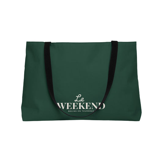A dark green tote bag with black shoulder straps and white printed text reading 'Le WEEKEND BEGINS ON THURSDAY' on a plain white background.
