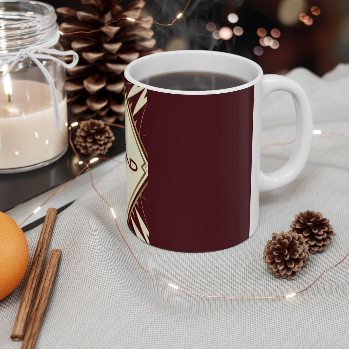 A white ceramic mug filled with a dark beverage, featuring a deep burgundy wraparound design with cream geometric patterns. The mug is styled on a light fabric surface with warm holiday props including pinecones, fairy lights, cinnamon sticks, a candle, and an orange.