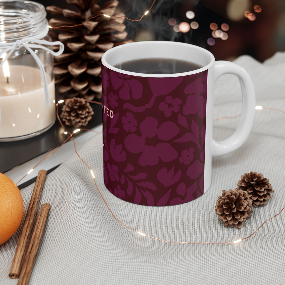 White mug with a dark purple accent on a table with decorative items including a candle, oranges, and pine cones.