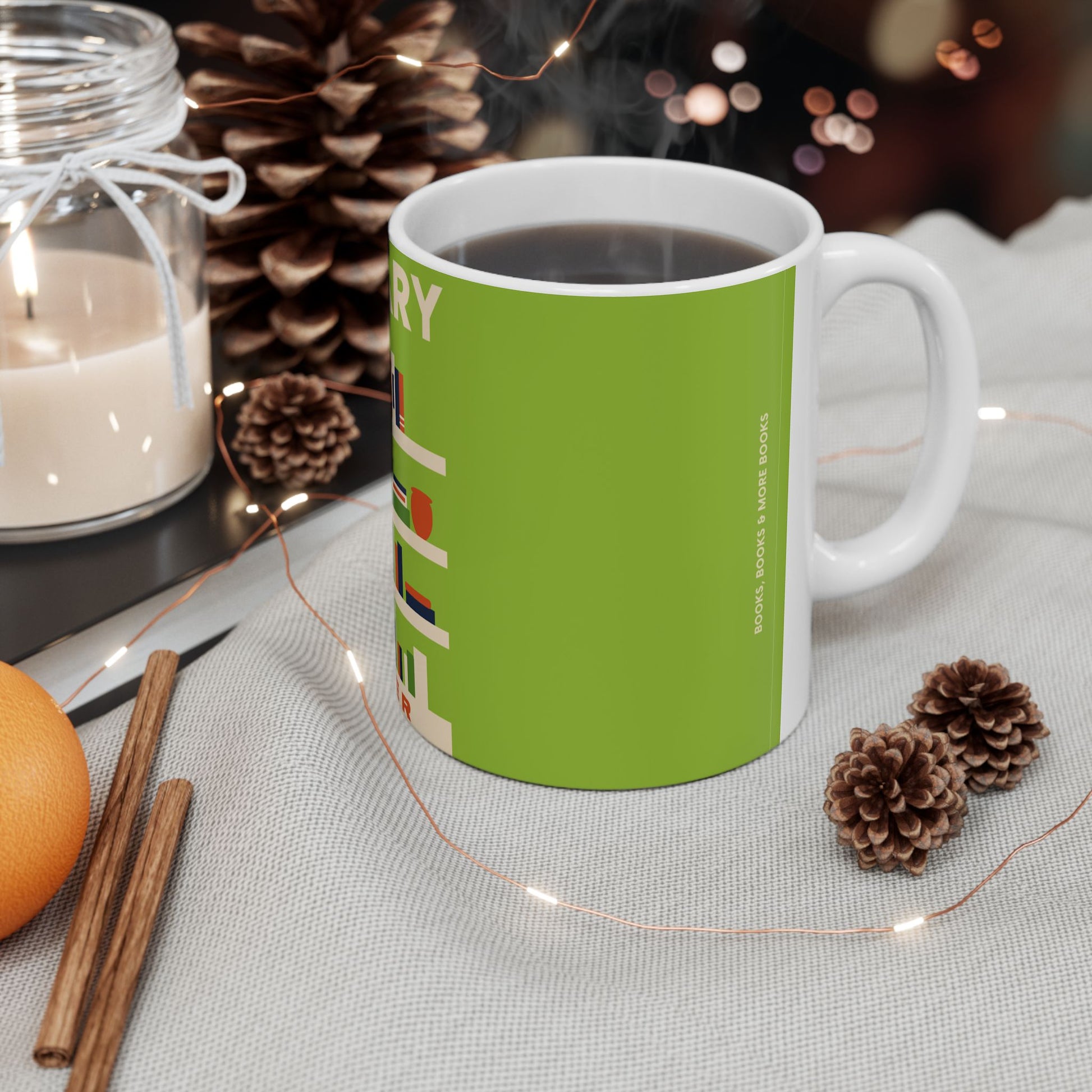 A white ceramic mug filled with a dark beverage, wrapped in a bright green sleeve featuring vertical text, styled on a light fabric surface with pinecones, a candle, cinnamon sticks, and fairy lights.