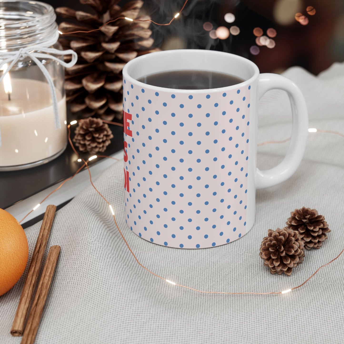 White mug with hot chocolate, surrounded by decorative elements like a candle, cinnamon sticks, and pine cones on a table.