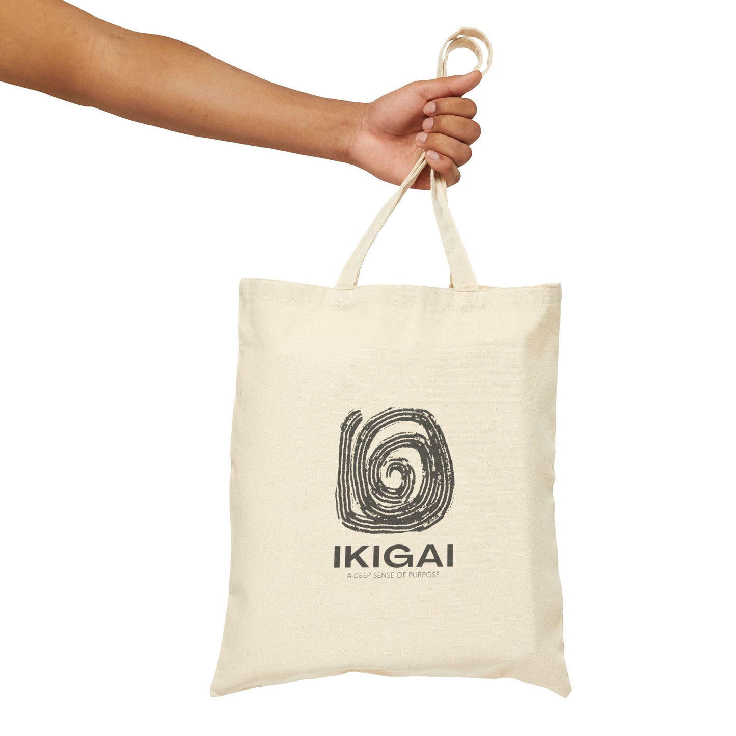 An off-white canvas tote bag with a black spiral graphic and the text 'IKIGAI' held by a person's hand against a solid white background.