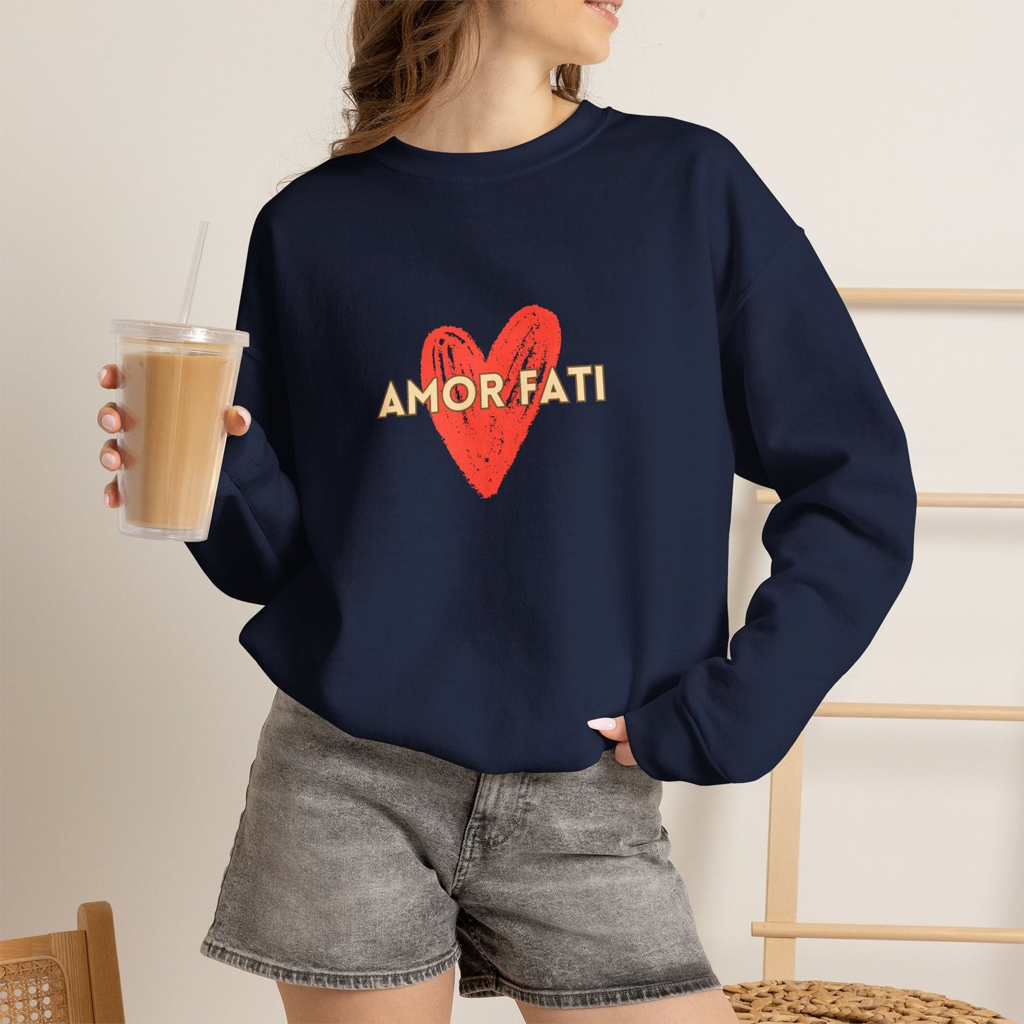 A woman wearing a navy blue crewneck sweatshirt with a red heart and the words 'AMOR FATI' printed on the front, paired with gray denim shorts and holding an iced coffee.