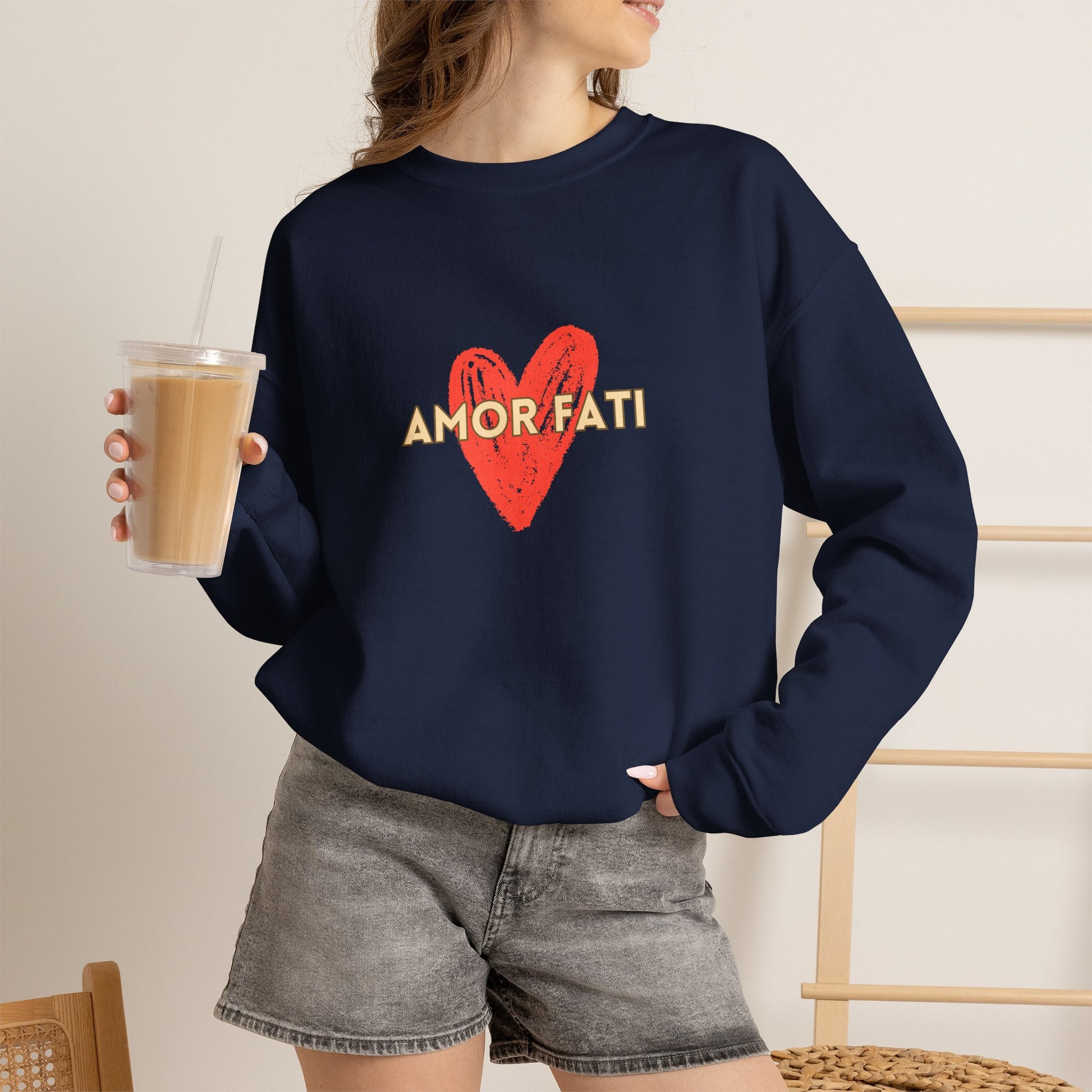 A woman wearing a navy blue crewneck sweatshirt with a red heart and the words 'AMOR FATI' printed on the front, paired with gray denim shorts and holding an iced coffee.