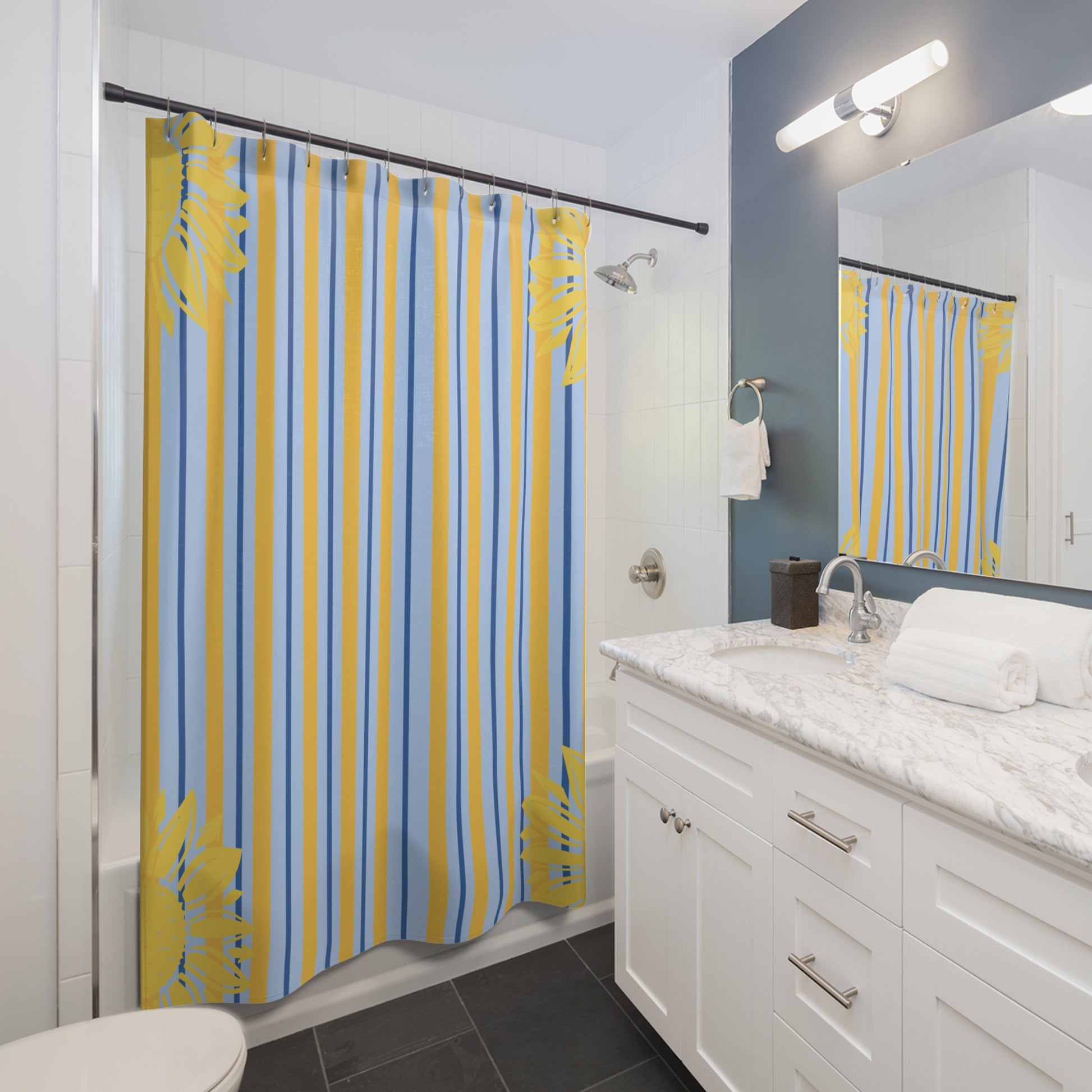 A bathroom shower curtain with vertical yellow and blue stripes and yellow tropical leaf motifs, hanging in a modern bathroom with a white vanity, marble countertop, and a mirror reflecting the curtain.