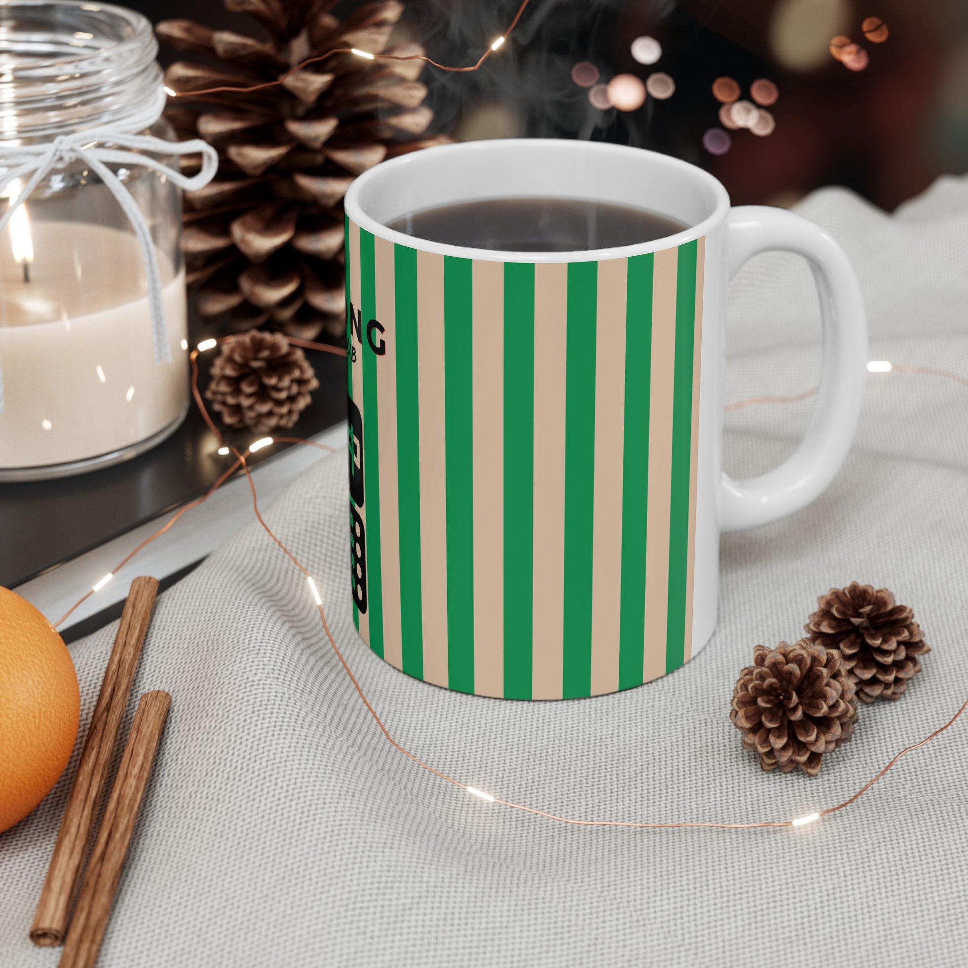 Green and beige striped mug with a candle, cinnamon stick, and orange on a table.