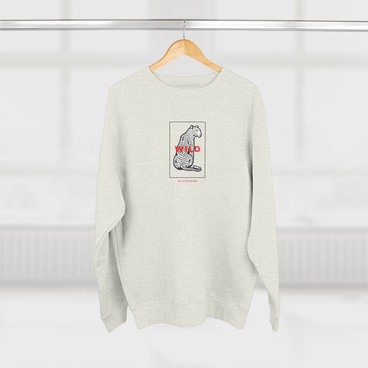 A light heather gray crewneck sweatshirt hanging on a wooden hanger against a bright, softly blurred window background. The sweatshirt features a central graphic of a black and white spotted cat inside a thin square border, with the word 'WILD' in red text and 'LIFE OF PEACH' printed below in small red letters.