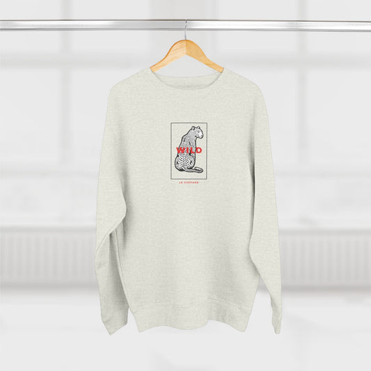 A light heather gray crewneck sweatshirt hanging on a wooden hanger against a bright, softly blurred window background. The sweatshirt features a central graphic of a black and white spotted cat inside a thin square border, with the word 'WILD' in red text and 'LIFE OF PEACH' printed below in small red letters.