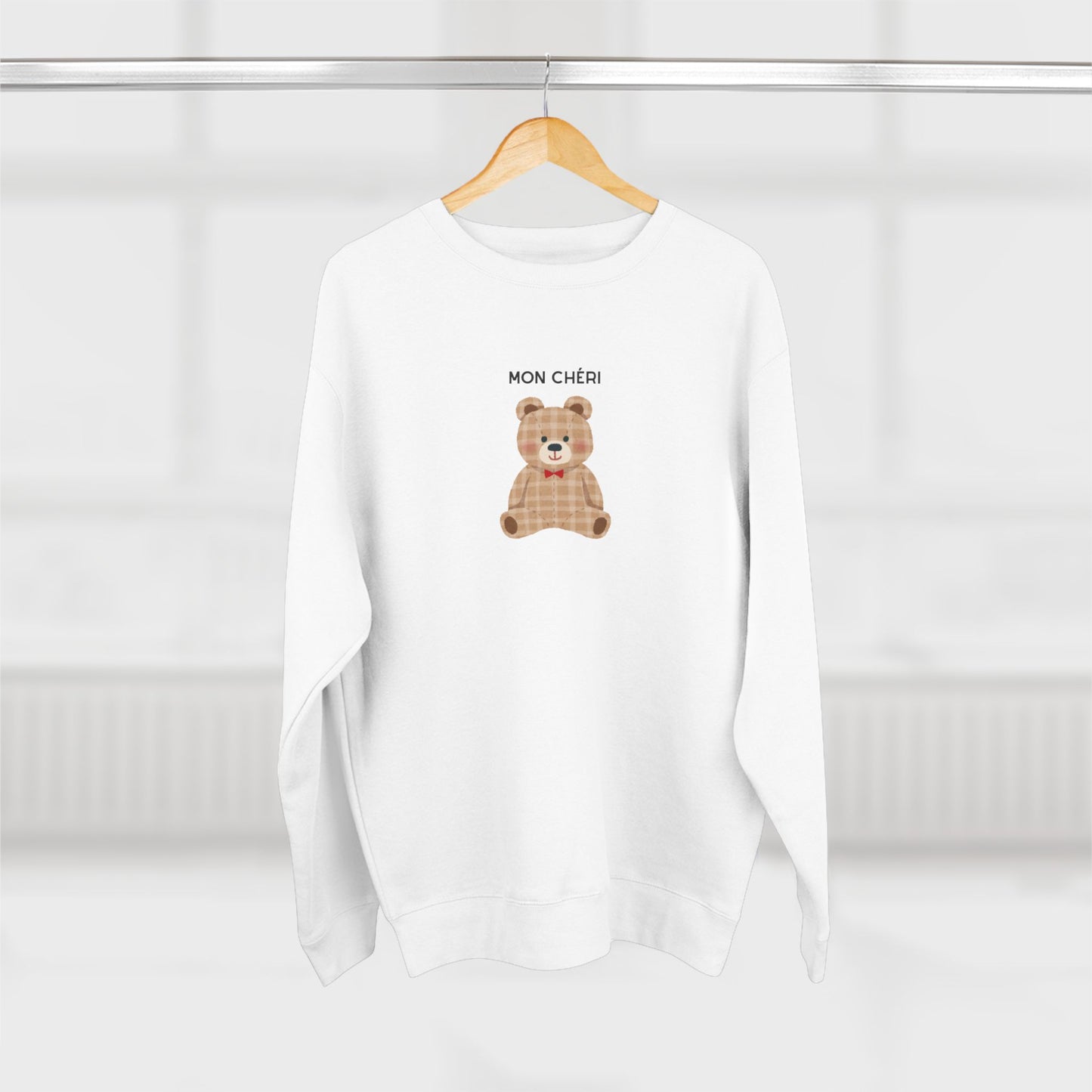White sweatshirt with a teddy bear graphic and 'MON CHÓRI' text on a hanger against a white background