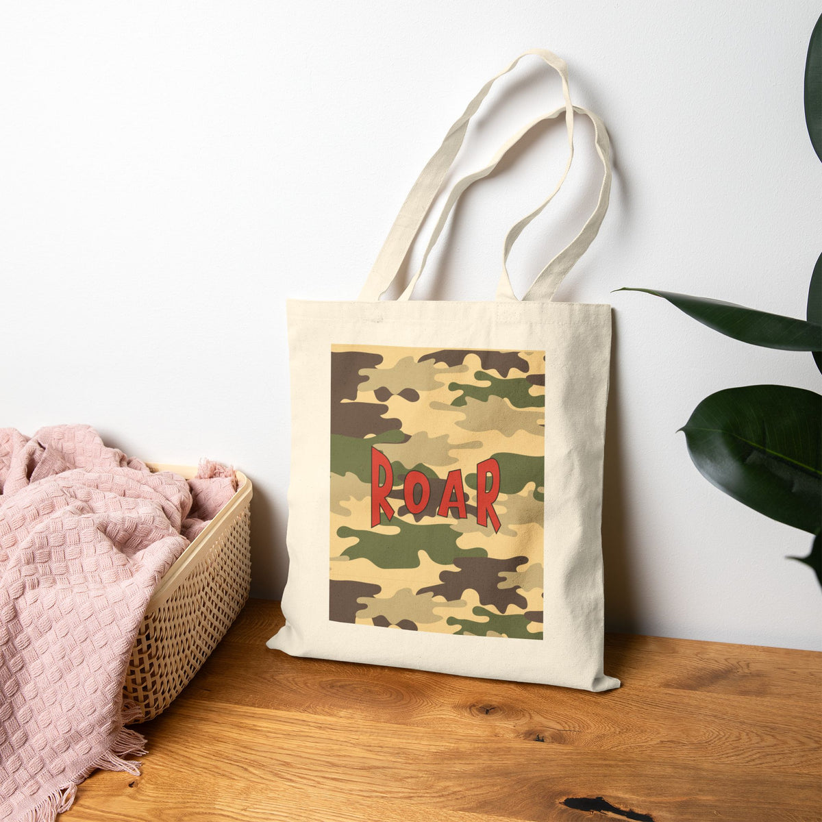 A canvas tote bag with a camouflage print and the word 'ROAR' in red letters, leaning on a wooden table next to a pink blanket and a green plant.