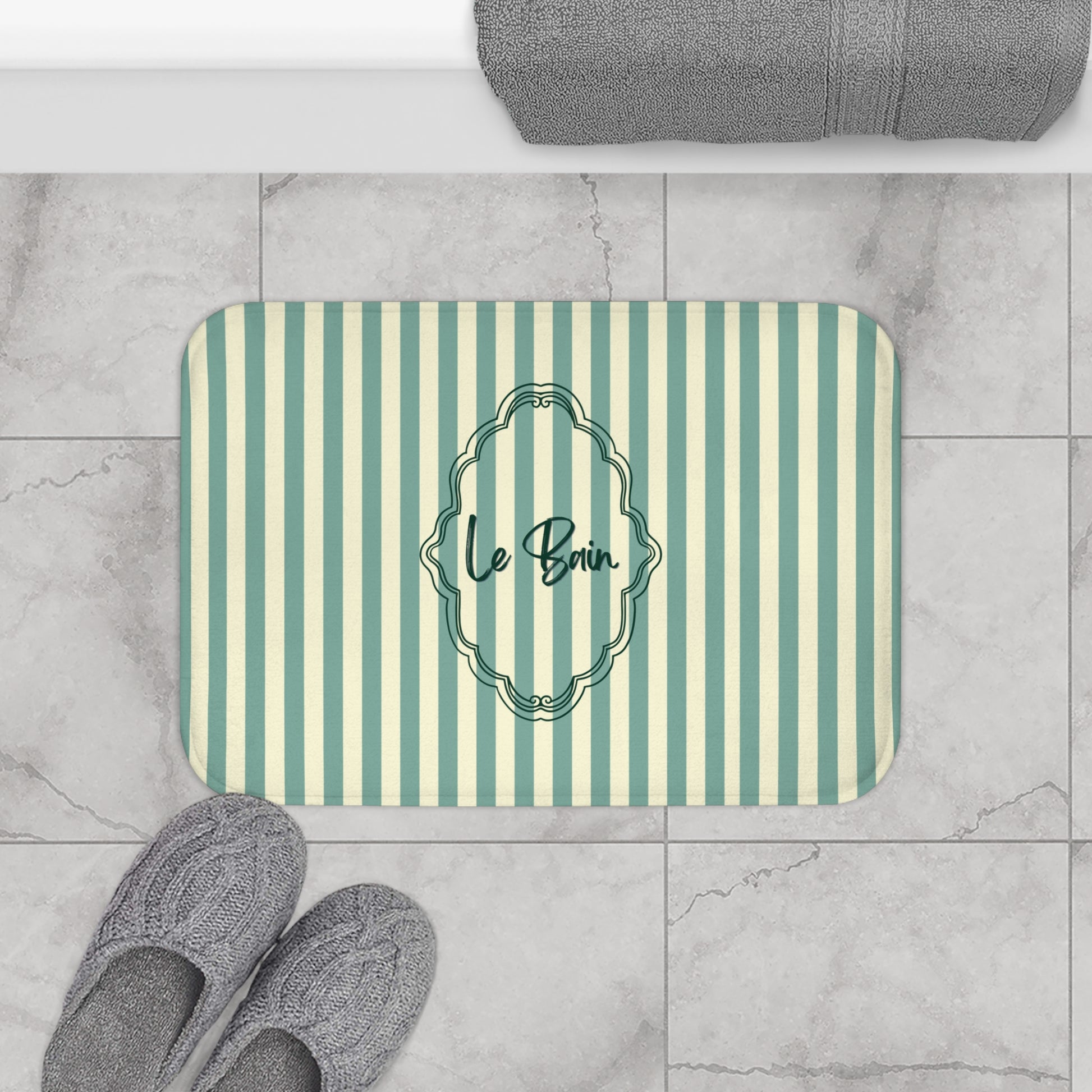 Striped bath mat with 'Le Bain' text on a tiled floor with slippers and a towel.