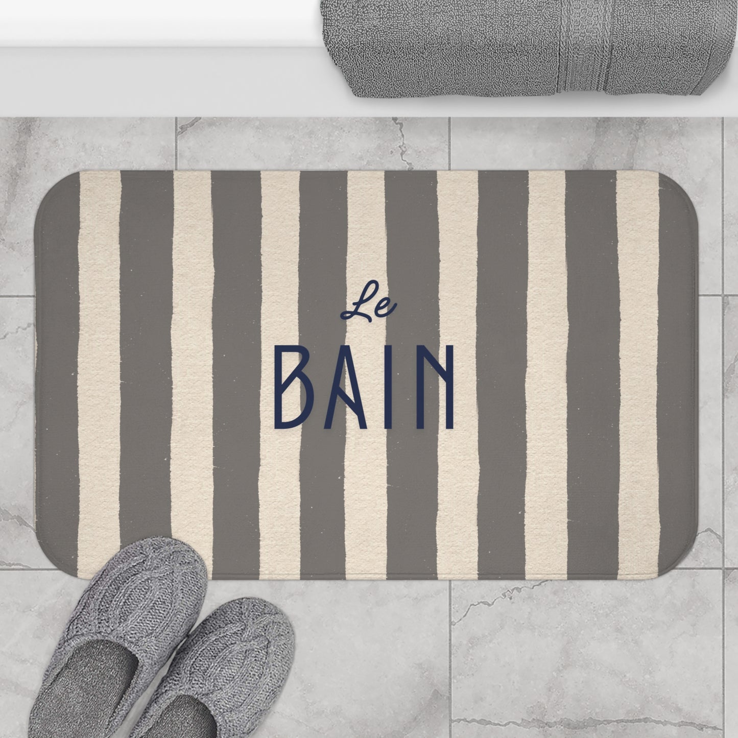 Striped bath mat with 'Le Bain' text on a marble floor with slippers and towel.