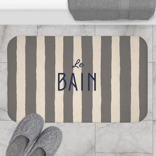 Striped bath mat with 'Le Bain' text on a marble floor with slippers and towel.