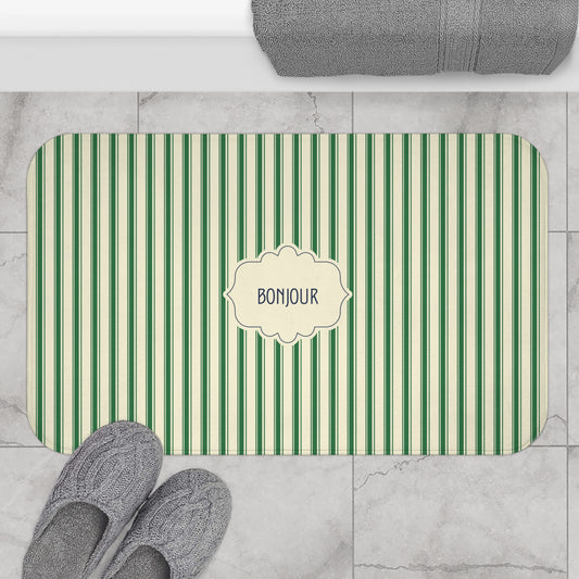 A rectangular bath mat with rounded corners, featuring vertical green and cream stripes and a central cream label that reads 'BONJOUR'. The mat is styled in a bathroom setting on a light gray marble tile floor, with gray slippers at the bottom left, a folded gray towel at the top, and the mat itself centered.